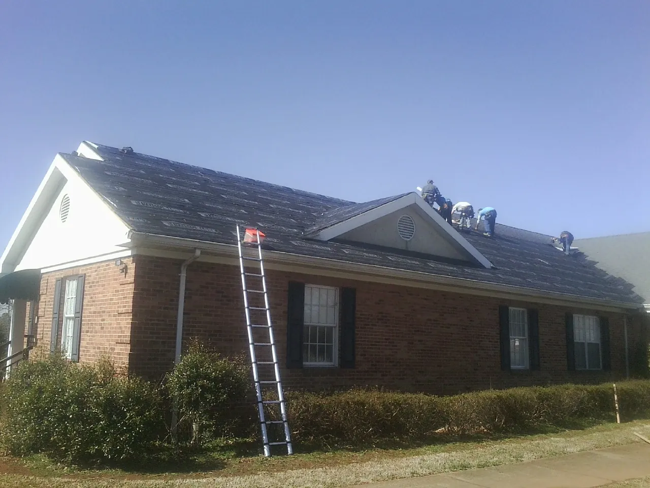 Crew tearing shingles off a brick ranch roof with a ladder set — Emergency Roof Repair work by our crew in Pontotoc