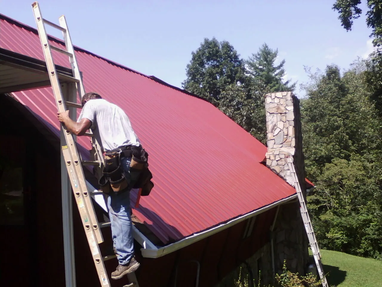 Roofer on a ladder beside a red metal roof and stone chimney — Soffit & Fascia Repair by Roofing Brothers in Pontotoc